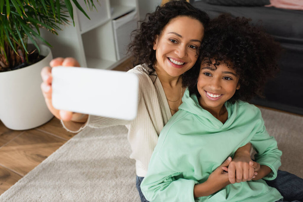 vista de ángulo alto de la madre afroamericana feliz tomando selfie con alegre hija preadolescente en el teléfono inteligente - Foto, Imagen