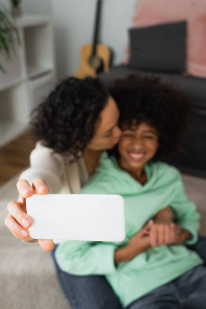 hoge hoek uitzicht van krullend Afrikaans Amerikaans moeder zoenen wang van dochter en het nemen van selfie op gerichte smartphone - Foto, afbeelding