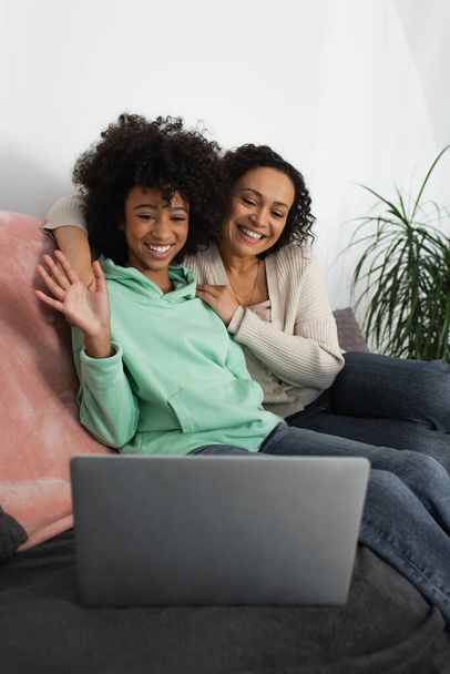 happy african american mother hugging cheerful preteen daughter waving hand during video call  - Foto, Imagem