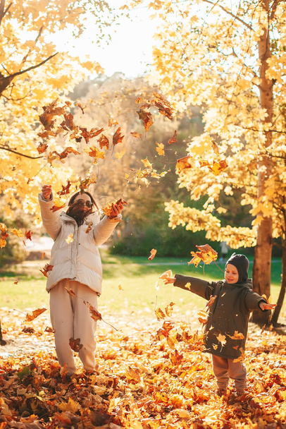 Happy mother with her little son throwing autumn leaves, playing in the autumn park. Autumn leaf fall. Family having fun together outside. - Foto, afbeelding