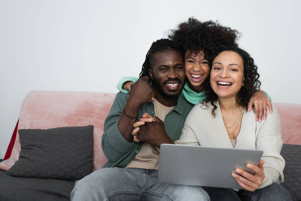 cheerful african american family smiling while looking at camera near laptop in living room  - Zdjęcie, obraz