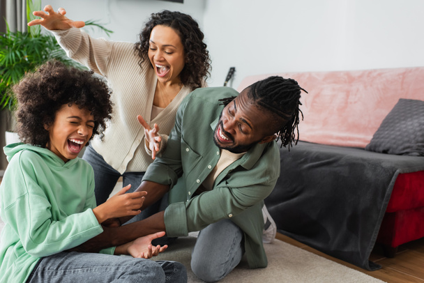 joyful african american father tickling preteen daughter near happy wife in living room  - Photo, Image