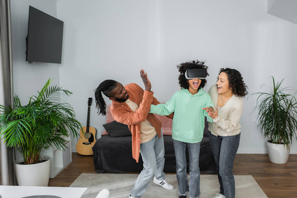 positive african american parents gesturing near preteen daughter gaming in vr headset in living room  - Fotografie, Obrázek