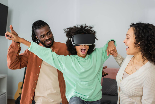 happy african american parents holding hands of amazed preteen daughter gaming in vr headset in living room  - Fotó, kép