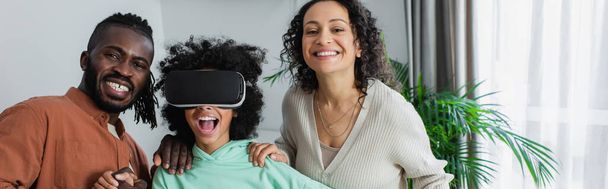 χαρούμενα αφρικανικής Αμερικής γονείς κοντά κατάπληκτος προ εφηβική κόρη gaming σε VR ακουστικά, πανό - Φωτογραφία, εικόνα
