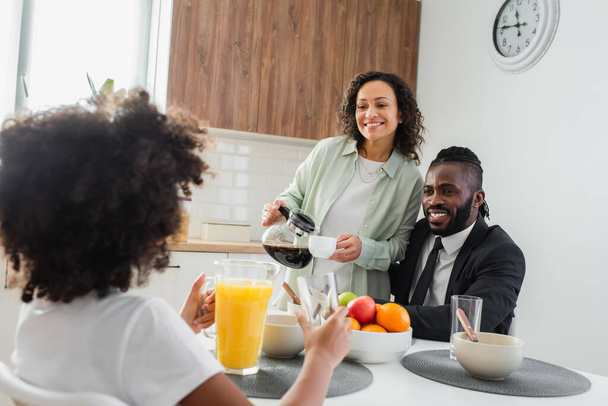 happy african american woman holding coffee pot and cup near husband in suit and preteen daughter during breakfast  - Foto, Imagem