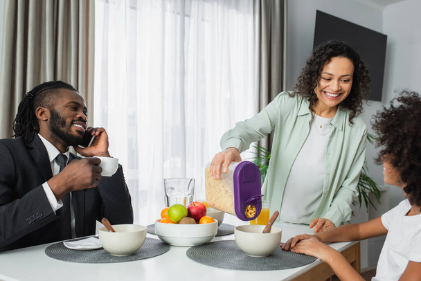 Веселая африканская американка, наливающая кукурузные хлопья в миску дочери рядом с мужем в костюме во время завтрака  - Фото, изображение