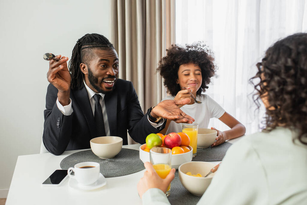 hombre afroamericano feliz en traje hablando con esposa cerca de hija preadolescente durante el desayuno  - Foto, imagen