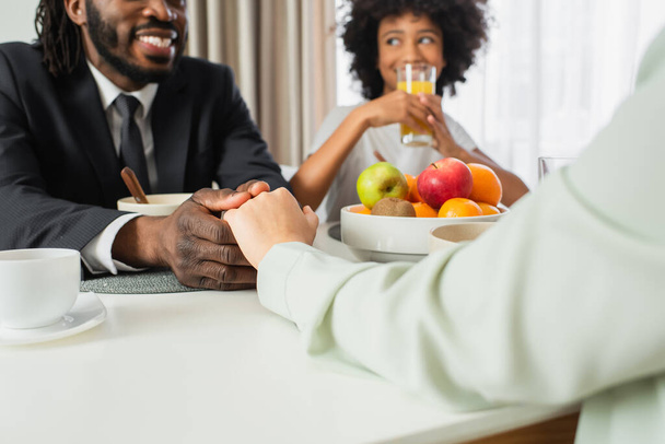 happy african american man in suit holding hands with wife near daughter during breakfast  - Photo, Image