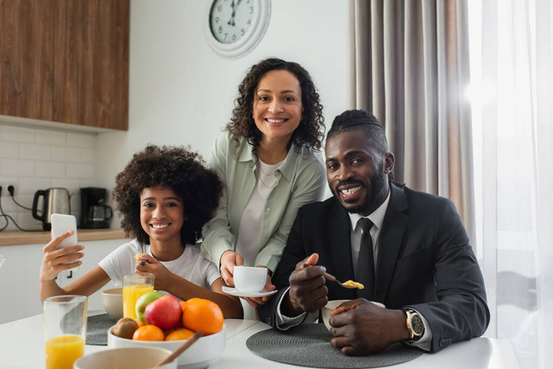 cheerful african american woman holding cup of coffee near husband in suit and preteen daughter with smartphone  - Valokuva, kuva