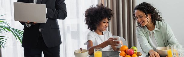 スーツ姿のアフリカ系アメリカ人男性がノートパソコンを持って立ち上がり幸せな娘が朝食中に母親にスマートフォンを見せながら - 写真・画像
