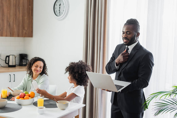 happy african american man in suit using laptop while girl showing smartphone to mother during breakfast  - Fotografie, Obrázek