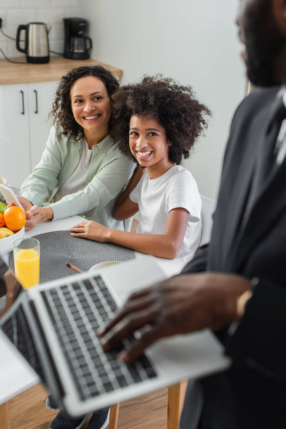 cheerful african american mother holding smartphone near daughter and looking at husband in suit using laptop on blurred foreground  - Zdjęcie, obraz