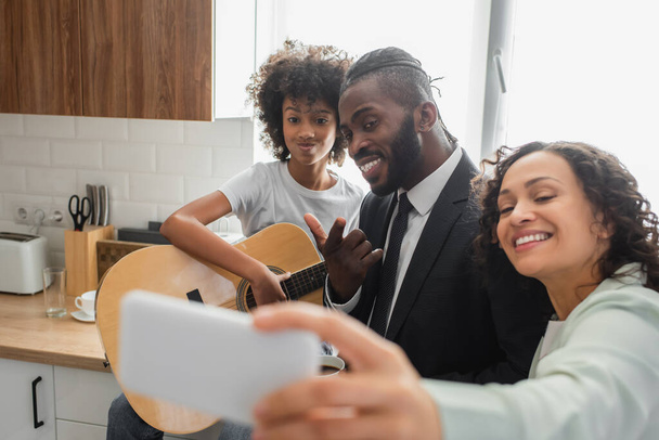 feliz afroamericana mujer tomando selfie con marido y preadolescente hija jugando guitarra acústica - Foto, Imagen
