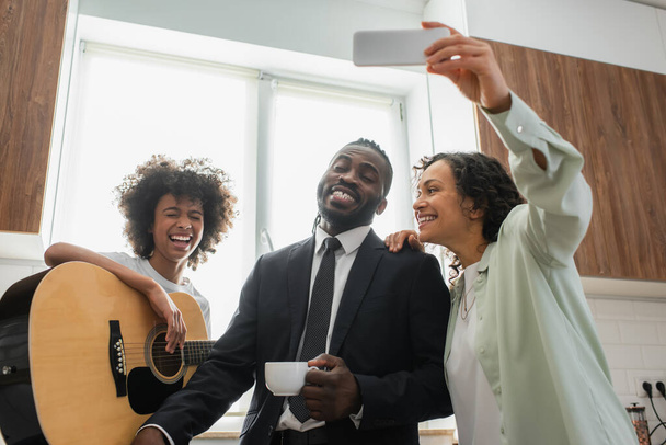 happy african american woman taking selfie with husband in suit and preteen daughter playing acoustic guitar - Photo, Image