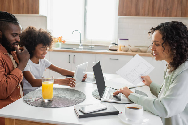 happy african american woman holding paper near laptop and family using digital tablet in kitchen  - Foto, Bild