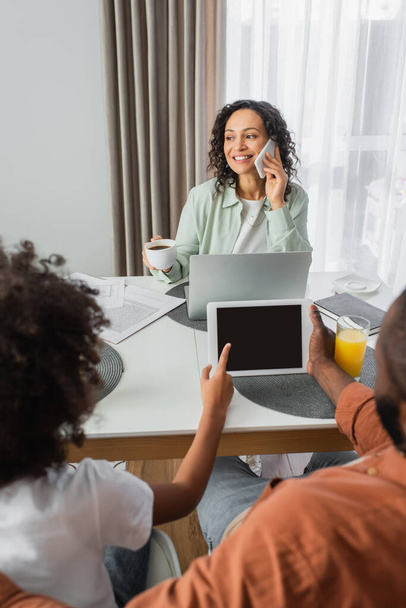heureuse femme afro-américaine parlant sur smartphone près d'un ordinateur portable tout mari et enfant en utilisant une tablette numérique dans la cuisine  - Photo, image
