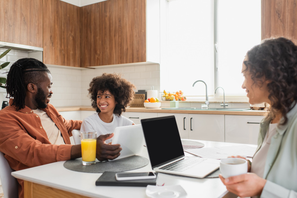happy african american woman holding cup near laptop while husband and daughter talking near digital tablet in kitchen  - Fotoğraf, Görsel
