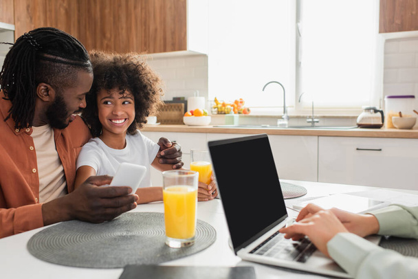 boldog afro-amerikai apa gazdaság smartphone és ölelés lánya, míg a feleség dolgozik otthonról laptop - Fotó, kép