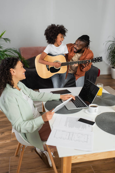 happy african american woman holding digital tablet while looking at daughter playing acoustic guitar near husband - Foto, Imagem