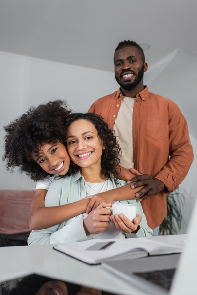 cheerful african american girl hugging mother with cup of coffee near smiling father - Foto, Imagem