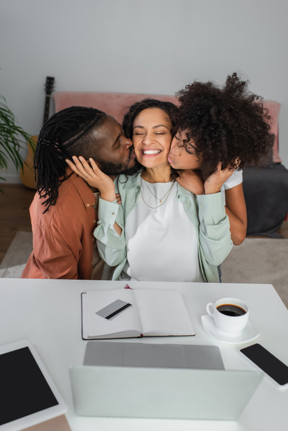 アフリカ系アメリカ人男性と10代の少女が机の上の装置の近くで幸せな女性の頬にキス - 写真・画像