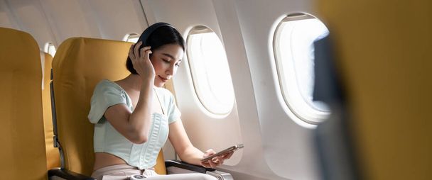 Young woman with mobile phone and headphones listening to music in airplane during flight. - Photo, Image