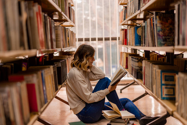 Ragazza che pensa e legge mentre è seduta in biblioteca tra gli scaffali - Foto, immagini
