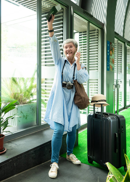 Ritratto di anziana anziana donna asiatica anni '60 sorridente in piedi con valigia bagaglio borsa e utilizzando smartphone per concetto di viaggio. - Foto, immagini