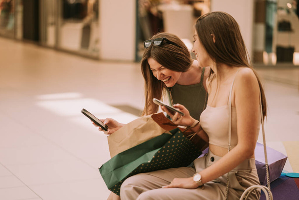 Two pretty girls laughing while sitting on the sofa in the mall - 写真・画像