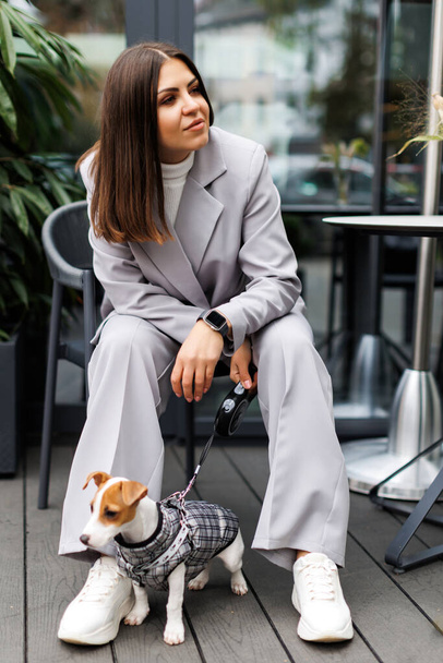 Woman in casual jacket sitting with her favorite fluffy trained purebred Jack Russell Terrier dog dressed in suit for dogs outdoors in cafe in autumn day. Faithful best friends - Photo, Image