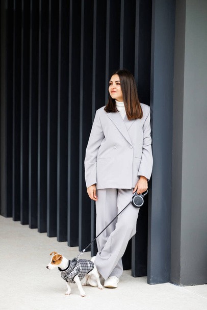 Woman in casual jacket standing with her favorite fluffy trained purebred Jack Russell Terrier dog dressed in suit for dogs near wall in outdoor in autumn day. Faithful best friends - Fotoğraf, Görsel