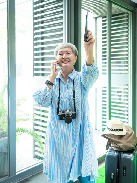 Portré idősebb felnőtt idős ázsiai nő 60-as évek mosolygós álló bőrönd bőrönd táska és az okostelefon utazási koncepció. - Fotó, kép