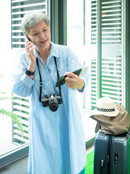 Portrait d'une femme asiatique âgée âgée des années 60 souriant debout avec sac à bagages valise et utilisant un smartphone pour concept de voyage. - Photo, image
