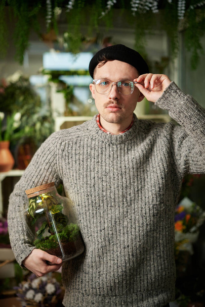 Atraktivní vousatý samec držící sklenici s rostlinou v květinářství. Dobře vypadající květinář ve svetru, beanie a brýlích. Malý obchodní koncept. Vysoce kvalitní vertikální obrázek - Fotografie, Obrázek