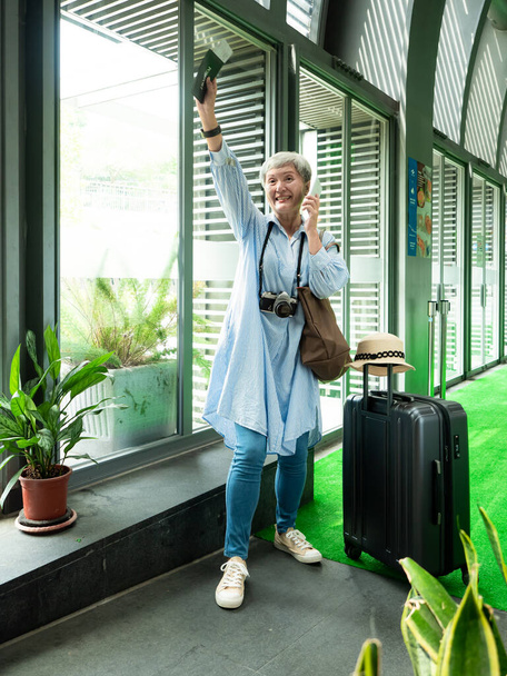 Портрет пожилой женщины 60 лет, улыбающейся стоя с чемоданом и используя смартфон для путешествий. - Фото, изображение