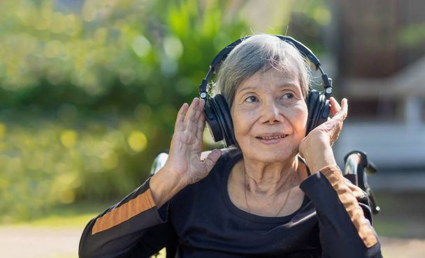 Ασιάτισσα ηλικιωμένη γυναίκα ακούει μουσική με ακουστικά στην πίσω αυλή. - Φωτογραφία, εικόνα