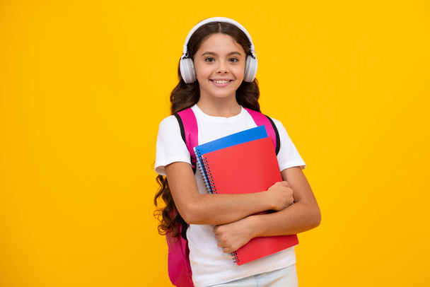 Okula dönelim. Sırt çantalı ve kulaklıklı liseli kız kitapları öğrenmeye hazır tutuyor. İzole edilmiş sarı stüdyo geçmişi olan okul çocukları. - Fotoğraf, Görsel