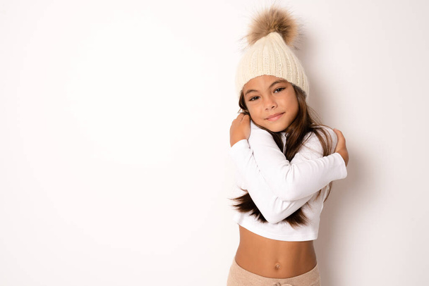 Roztomilé usmívající se dívka na sobě zimní klobouk objímající se na bílém pozadí - Fotografie, Obrázek