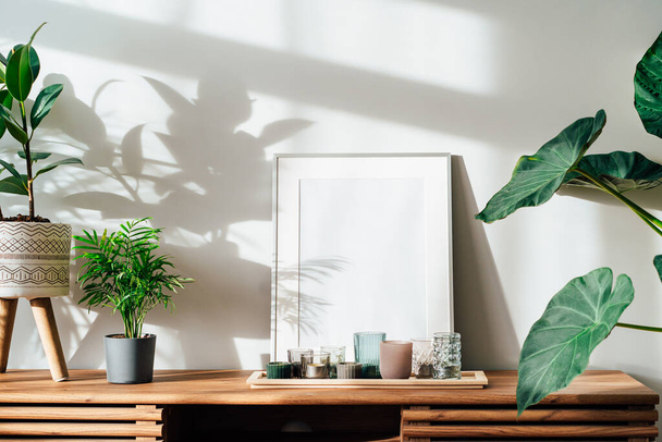 Modernes minimalistisches Interieur im skandinavischen Stil mit weißen Poster-Attrappen, Kerzen und tropisch grünen Pflanzen auf einer Holzkonsole unter Sonnenlicht und Schatten an einer grauen Wand. Selektiver Fokus. - Foto, Bild