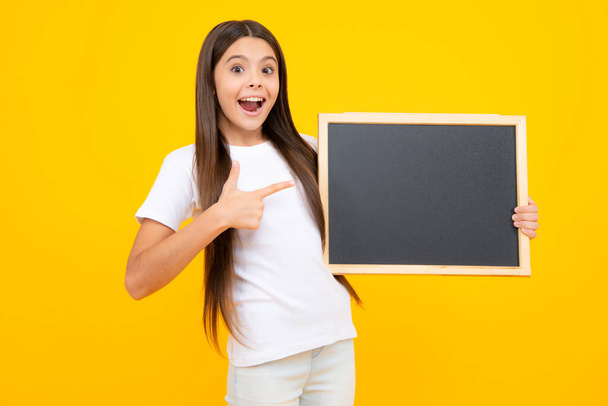 Teenager younf school girl holding school empty blackboard isolated on yellow background. Portrait of a teen female student. Excited teenager, glad amazed and overjoyed emotions - Photo, image