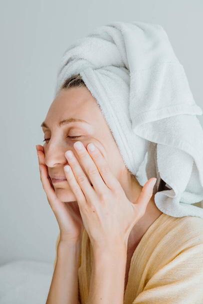 Jeune femme en peignoir et une serviette sur sa tête applique une crème sur sa peau, masse son visage et son corps. concept d'autosoin. - Photo, image