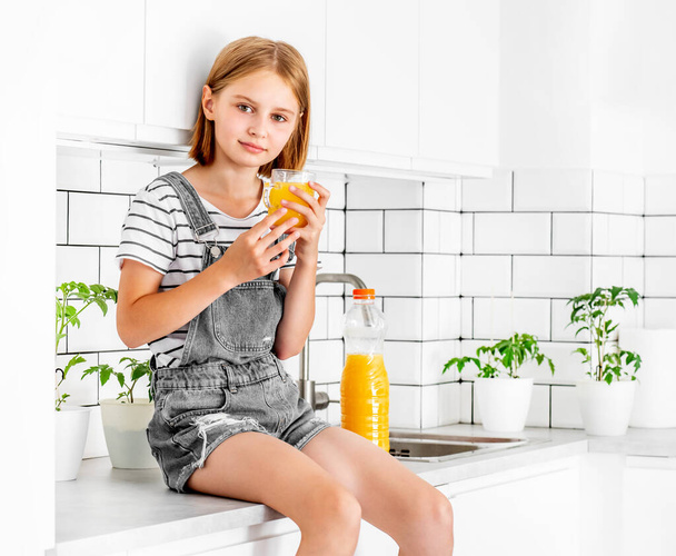Preteen girl with orange juice bottle and glass sitting at kitchen table. Pretty child kid with vitamin fruit drink at home - Foto, Imagen