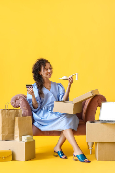 Young African-American woman with credit card and new shoes sitting in armchair on yellow background. Online shopping - Photo, Image