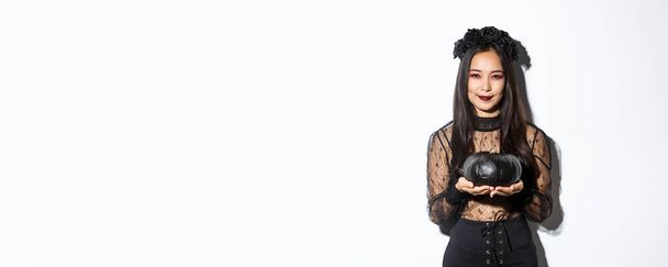 Улыбающаяся коварная азиатка в костюме ведьмы, празднующая Хэллоуин, держащая черную тыкву, стоящая на белом фоне. - Фото, изображение