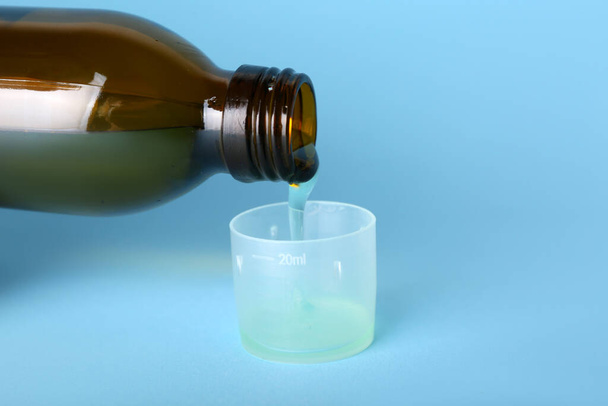 Ρίχνουμε σιρόπι για το βήχα από το μπουκάλι στο φλιτζάνι σε μπλε φόντο - Φωτογραφία, εικόνα