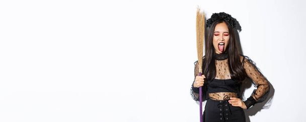 Beztroska zuchwała Azjatka ciesząca się imprezą halloween, nosząca kostium czarownicy i trzymająca miotłę, pokazująca język, stojąca nad białym tłem. - Zdjęcie, obraz