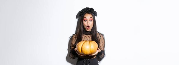 Immagine di stupita ragazza asiatica guardando grande zucca affascinato, indossando abito gotico e ghirlanda nera, in piedi su sfondo bianco. - Foto, immagini
