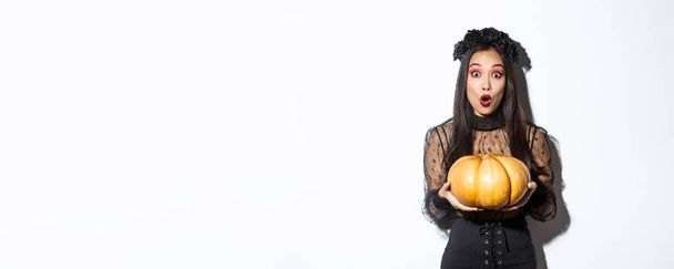 Sorprendida chica asiática en traje de bruja, sosteniendo la calabaza y jadeando asombrada ante la cámara, preparándose para las vacaciones de Halloween, de pie sobre fondo blanco. - Foto, Imagen
