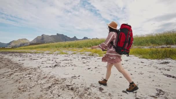 A tourist girl with a big red backpack and a hat walks along the seashore in the mountains. beautiful norway summer landscape - Footage, Video